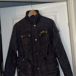 Svart quiltad jacka Barbour International - Quiltad svart jacka från Barbour International med guldiga knappar, hög krage och bälte i midjan. Jackan har flera fickor framtill och klassisk Barbour-logga på bröstet. Perfekt för dig som vill ha en stilren och tidlös look.