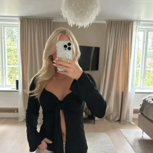 Svart omlottopp med knyt framtill - Säljer en svart långärmad omlottopp med snygg knytning framtill och djup v-ringning. Toppen har en croppad passform och långa ärmar, perfekt för att skapa en trendig look. Materialet känns mjukt och följsamt mot kroppen.