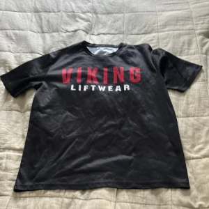 Viking liftwear gymtröja - Svart t-shirt från Viking Liftwear med röd och vit text på framsidan och en stor vikingahjälm i vitt och rött på ryggen. T-shirten har rund hals och korta ärmar, perfekt för gymmet eller chill. Materialet känns som polyester och har en loose passform.