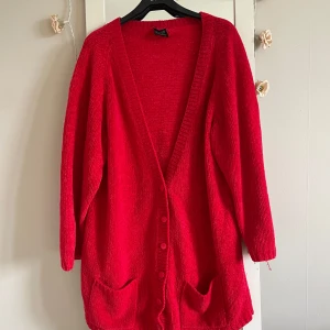 Röd stickad kofta från Carling - Snygg röd stickad kofta från Carling med lång ärm, djup v-ringning och knappar framtill. Modellen är loose fit och har två praktiska fickor framtill. Perfekt för dig som gillar färg och vill ha en mysig, oversized look.