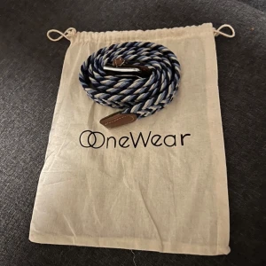  Flätat bälte från OneWear - Snyggt flätat bälte från OneWear i blått, vitt. Bältet har ett randigt mönster och detaljer i brunt skinn vid spännet. Perfekt bälte för att lyfta din outfit med en klassisk och stilren look.