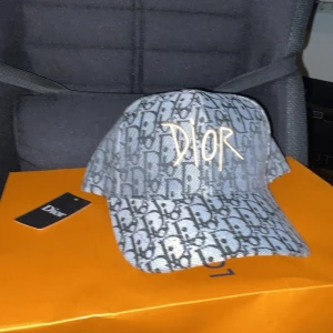 Dior keps  - Säljer en snygg keps från Dior med klassiskt monogrammönster i svart och grått. Kepsen har broderad Dior-logga framtill i vitt och är tillverkad i ett slitstarkt tygmaterial. Perfekt accessoar för att lyfta din outfit.