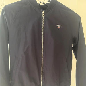 Mörkblå bomberjacka från GANT - Snygg mörkblå bomberjacka från GANT med klassisk siluett och ribbade muddar. Jackan har dragkedja framtill, två sidofickor och GANT-logga broderad på bröstet. Perfekt för dig som gillar stilrena och sportiga plagg.