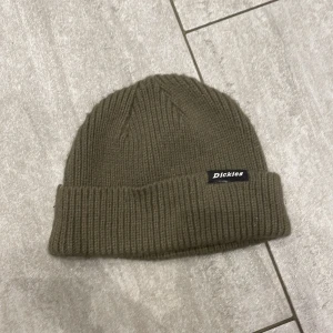 Olivgrön ribbstickad mössa från Dickies - Snygg olivgrön mössa från Dickies med ribbstickad design och uppvikt kant. Klassisk och enkel stil med liten svart logotyp på kanten. Perfekt för kalla dagar och passar till de flesta outfits.