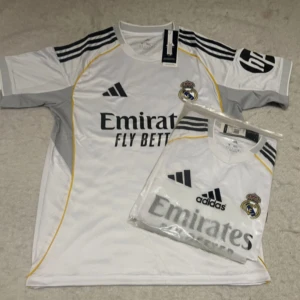 Real Madrid hemmatröja Adidas vit - Snygg Real Madrid hemmatröja från Adidas i vitt med svarta detaljer och gula inslag. Tröjan har klubbmärke på bröstet, sponsortryck och klassiska Adidas-ränder på axlarna. Tillverkad i lätt och ventilerande material, perfekt för match eller träning.