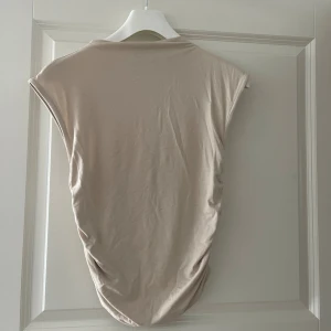 Beige ärmlös linnetopp - Säljer en stilren beige linnetopp med ärmlös design och lätt drapering i sidorna. Toppen har en enkel rund halsringning och är tillverkad i ett mjukt, stretchigt material som känns skönt mot huden. Perfekt att matcha med jeans eller kjol för en clean look.
