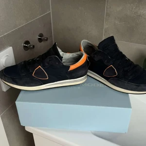 Svarta sneakers med orange detalj - Snygga svarta sneakers från Philippe Model med orange detalj vid hälen och på sidan. Skorna har snörning, vit sula och är tillverkade i mocka och textil. Perfekta för dig som gillar en sportig och trendig look.