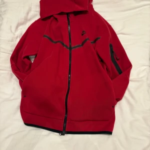 Röd Nike vindjacka med huva M - Röd vindjacka från Nike med svart dragkedja, huva och svarta detaljer. Jackan har en sportig look med en svart rand över bröstet och en ficka med dragkedja på ärmen. Perfekt för dig som gillar streetwear och sportig stil. Byxor ingår!! Finns även i grön