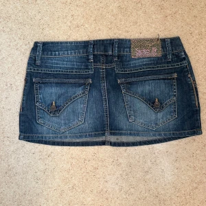 Jeanskjol låg midjad  - Snygg kort jeanskjol  i klassisk blå denim. Kjolen har fem fickor, coola bakfickor med lock och knapp, samt bälteshällor. Midjan mäter ca 40 cm. Perfekt för dig som gillar en avslappnad och trendig stil.