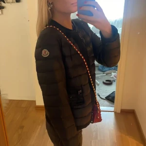 Svart Moncler pufferjacka dam - Snygg svart pufferjacka från Moncler med klassisk logga på ärmen. Jackan har dragkedja framtill, två fickor med dragkedja och en stilren, quiltad design. Perfekt för kyliga dagar och ger en clean look till alla outfits.