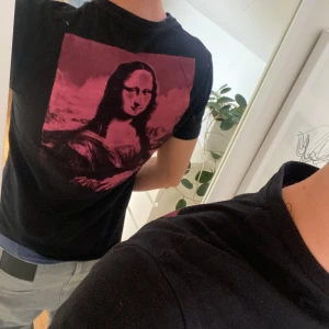 Svart t-shirt med rosa Mona Lisa-tryck - Svart t-shirt från Yourturn med stort rosa Mona Lisa-tryck på ryggen och mindre rosa tryck med ögon och text på bröstet. Klassisk passform och rund hals. Skön bomullskänsla och riktigt snygg streetstil.