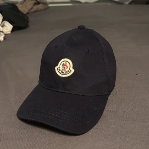 Svart keps från Moncler - Stilren svart keps från Moncler med klassisk böjd skärm och justerbart spänne bak. Framtill sitter Monclers ikoniska broderade logotyp i vitt, rött och blått. Kepsen är tillverkad i bomull och passar perfekt för en clean och sportig look.