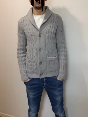 Cardigan - Säljer en ljusgrå kabelstick matte kofta med stora knappar framtill och två fickor. Koftan har en bred krage och är långärmad, perfekt för kyligare dagar. Snygg och enkel att matcha till olika outfits.