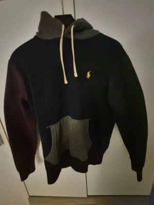 Polo ralph lauren hoodie - Snygg och bekväm hoodie från Polo Ralph Lauren i en unik färgkombination. Tröjan har en mörkblå bas, med ena ärmen i grönt och den andra i rött, vilket ger ett sportigt och distinkt uttryck. Huva och magficka i grått skapar en snygg kontrast och förstärker den moderna designen. På bröstet finns den klassiska broderade Polo-loggan i gult. Tillverkad i mjukt och värmande material som passar perfekt för både vardag och fritid. En statement-hoodie för dig som vill sticka ut med stil.