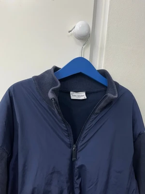 Marinblå bomberjacka från John Henric - Säljer en marinblå bomberjacka från John Henric. Jackan har ribbade muddar vid krage, ärmslut och nederkant samt dragkedja framtill och på fickorna. Materialet är en mix av polyester och stickad bomull vilket gör den både lätt och bekväm.