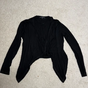 Svart kofta från Ilse Jacobsen - Svart tunn kofta från Ilse Jacobsen i storlek S. Koftan har lång ärm och öppen framtill med asymmetrisk nederkant som ger en cool och avslappnad look.