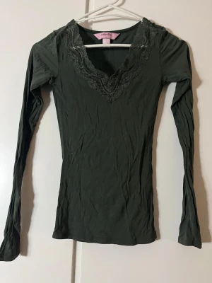 Mörkgrön spetsdetaljerad topp Hunkemöller - Supersnygg mörkgrön långärmad topp från Hunkemöller i stretchigt material. V-ringad med fina spetsdetaljer runt halsen som ger en romantisk vibe. Perfekt att styla med jeans eller kjol för en trendig look.