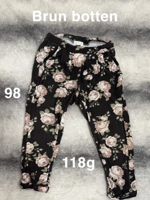 Blommiga byxor med brun botten - Mysiga byxor med brun botten och stora blommor i rosa och grönt. Byxorna har resår i midjan, perfekta för en avslappnad och trendig look. 🌸 Fint använt skick. Brun botten. Ytludd. Storlek 98 