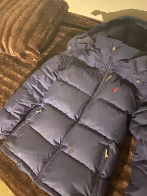 Mörkblå dunjacka från Polo Ralph Lauren - Säljer en mörkblå dunjacka från Polo Ralph Lauren med huva och dragkedjor. Jackan har det klassiska röda Polo-loggan broderad på bröstet och två sidofickor med dragkedja. Perfekt för kalla dagar och riktigt snygg passform. Du kan även kontakta mig för ytterligare bilder eller information. 