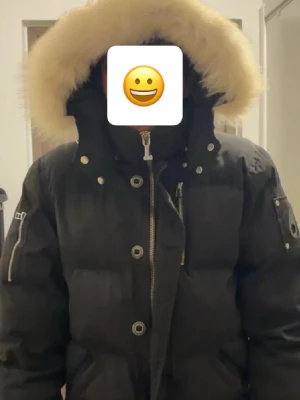 Svart dunjacka med pälskantad huva - Säljer en svart dunjacka från Moose Knuckles med stor fluffig pälskantad huva. Jackan har dragkedja och knappar framtill, samt detaljerade fickor och logga på ärmen. Perfekt för kalla dagar och riktigt snygg streetstil.