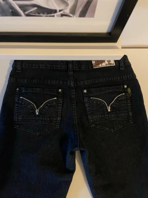 Svarta jeans med dragkedjor i bak - Unika svarta jeans med coola dragkedjor på bakfickorna och dekorativa stjärnformade nitar vid fickorna. Byxorna har rak passform och klassisk midja. Perfekt för dig som vill sticka ut med detaljer och snygg struktur. Aldrig använda, men köpt secondhand.