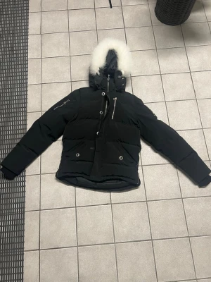  Moose Knuckles jacka S - Säljer en svart vinterjacka från Moose Knuckles med vit päls runt huvan och flera dragkedjor. Jackan har broderad logga på ärmen och coolt broderi på insidan. Perfekt för kalla dagar med sin tjocka dunfyllning och snygga detaljer.