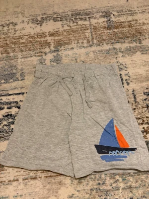 Grå shorts med segelbåt från H&M - Grå mjuka shorts från H&M med snörning i midjan och ett färgglatt tryck av en segelbåt i blått och orange på ena benet. Tillverkade i bomullsmix för skön känsla, perfekta för sommardagar.