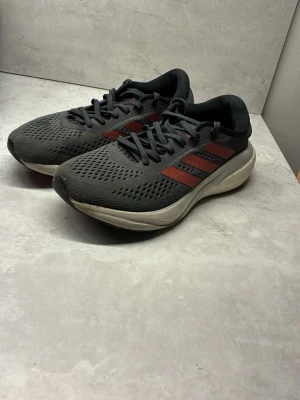 Adidas Supernova grå/röda sneakers - Säljer ett par Adidas Supernova sneakers i grått med röda ränder och vit sula. Ovandelen är i ventilerande syntetmaterial och skorna har klassisk snörning. Perfekta för dig som gillar sportig stil och vill ha bekväma skor till vardags.