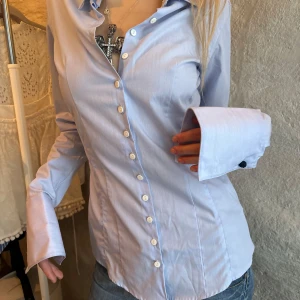 Figursydd skjorta - En ljusblå skjorta med klassisk krage och vita knappar hela vägen ner. Skjortan har långa ärmar med breda manschetter och är figursydd för en snygg passform. Perfekt till jeans eller kjol för en clean look.