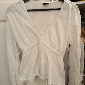 Vit topp från Gina Tricot - Säljer en vit topp från Gina Tricot i storlek M. Toppen har långa ärmar, v-ringning och knytband i midjan för en snygg passform. Tillverkad i ett lätt och mjukt bomullsliknande material som känns luftigt och fräscht.