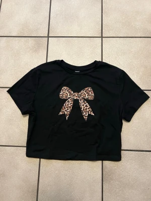 Svart crop t-shirt med leopardrosett - Svart croppad t-shirt med ett stort tryck av en rosett i leopardmönster på bröstet. T-shirten har korta ärmar och rund hals. Materialet är en mix av polyester, bomull och elastan vilket gör den mjuk och stretchig.