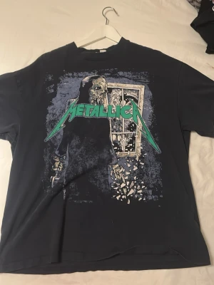 Svart Metallica t-shirt från H&M Divided - Säljer en svart t-shirt från H&M Divided med stor Metallica-print framtill. Trycket har en cool dödskalle och fönsterdesign i blått, vitt och grönt. Klassisk rund hals och korta ärmar. Perfekt för dig som gillar bandmerch och vill sticka ut. OVERSIZE så den är egentligen typ L 