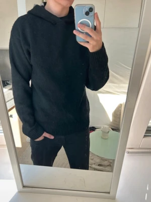 Svart hoodie i stickat material - Mysig svart hoodie i stickat material med huva och lång ärm. Tröjan har en loose passform och är perfekt för kyligare dagar. Enkel och stilren design utan tryck eller detaljer, passar dig som gillar en clean look.