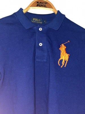 Blå långärmad henley från Polo Ralph Lauren - Säljer en blå långärmad henley från Polo Ralph Lauren med klassisk krage och två knappar framtill. Stor orange broderad logga på bröstet. Skjortan är i mjuk bomull och har en clean, tidlös look som funkar till allt.