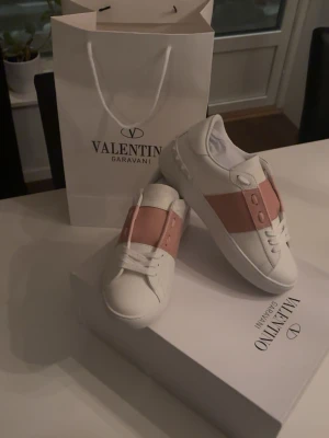 Valentino Garavani Open sneakers vit/rosa - Snygga Valentino Garavani Open sneakers i vitt skinn med bred rosa läderdetalj över vristen. Klassisk rund tå, vita snören och diskret logga på plösen. Sulan har ett rutigt mönster undertill och nitar baktill för extra edge. Perfekta för dig som gillar exklusiv streetstyle. (Fick i julklapp men var en storlek förliten, bara provade inomhus)  