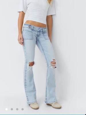 Ljusblå bootcut jeans med slitningar - Säljer ett par ljusblå bootcut jeans med låg midja och slitna detaljer över knäna. Jeansen har stora framfickor, dubbla knappar i linningen och är tillverkade i klassiskt denimtyg. Perfekta för en avslappnad och trendig look. Aldrig använda💕