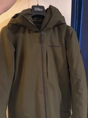 Grön vindjacka från Peak Performance - Säljer en grön skidjacka från Peak Performance med huva och hel dragkedja framtill. Jackan har en stilren design, diskret logga på bröstet och är tillverkad i slitstarkt syntetmaterial. Perfekt för kyliga dagar och outdoor-äventyr.
