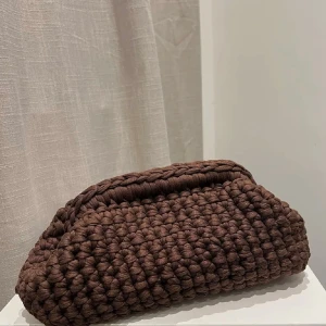 Brun virkad clutch väska - Säljer en virkad clutch i mörkbrunt. Väskan har en mjuk, handgjord känsla med grova maskor och en enkel avlång form. Perfekt för dig som vill ha en unik accessoar med cozy vibes. Den har även ett litet handtag som gör de lätt att hålla den🩷