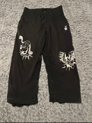 Svarta wide byxor från Harlaut Apparel - Unika svarta byxor från Harlaut Apparel med bred passform och vita grafiska tryck på både fram- och baksida. Byxorna har raka ben, bälteshällor och coola detaljer som ger streetwear-vibe. Perfekta för dig som vill sticka ut med din stil. Pris är inte satt i sten!