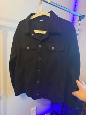 Svart jeansjacka med knappar - Säljer en svart jeansjacka med klassisk krage och metallknappar framtill. Jackan har två bröstfickor med lock och knapp, samt långa ärmar. Perfekt för dig som gillar en enkel och stilren look.