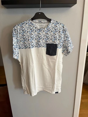  t-shirt från Tom Tailor - Snygg t-shirt från Tom Tailor med vit nederdel och överdel i blått, svart och vitt abstrakt mönster. Har en mörkblå bröstficka och rund hals. Tillverkad i ekologisk bomull, perfekt för dig som gillar stilrena detaljer och lite färg. Helt ny med lapp . Standard fit 