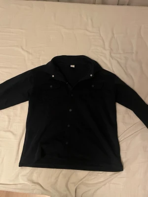 Svart overshirt från Lindex kanska mjuk matrial - Säljer en svart overshirt från Lindex i storlek L. Jackan har klassisk krage, tryckknappar framtill och två bröstfickor med lock. Perfekt lager-på-lager-plagg och enkel att styla till olika outfits.