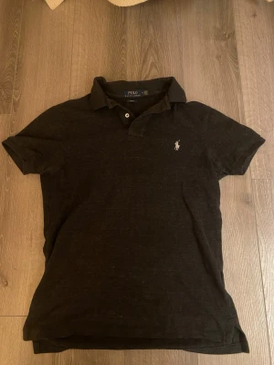 Svart piké från Polo Ralph Lauren - Klassisk svart pikétröja från Polo Ralph Lauren i slim fit. Tröjan har korta ärmar, krage, två knappar och den ikoniska broderade loggan på bröstet. Tillverkad i mjuk bomull som känns skön mot huden.