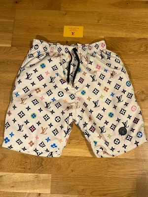 Louis Vuitton vita mönstrade badshorts  - Louis Vuitton shorts i vitt med ikoniskt monogrammönster i flera färger. Elastisk midja med svart snörning för skön passform. Perfekta för sommaren och ger en lyxig vibe till din outfit.