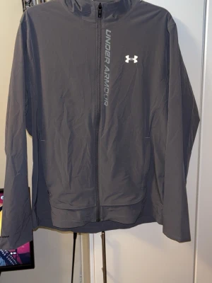 Grå vindjacka från Under Armour - Säljer en grå vindjacka från Under Armour med huva och dragkedja framtill. Jackan har logga och text på bröstet, samt ventilerande meshfoder på insidan. Perfekt för träning utomhus när det blåser. Snygg och sportig look!