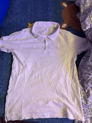 Vit piké från Lindex med brodyr - Klassisk vit pikétröja från Lindex med krage och två knappar framtill. Diskret broderad logga på bröstet. Tillverkad i mjuk bomull som känns skön mot huden. Perfekt till jeans eller shorts för en clean look.