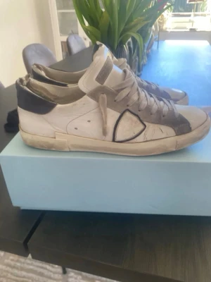 Philippe Model vita sneakers med grå detaljer - Säljer ett par Philippe Model sneakers i vitt med grå och svarta detaljer. Skorna har snörning, rund tå och platt sula. Materialet är skinn och mocka, och de kommer med originalkartong och dustbag. Perfekt för dig som gillar stilrena och exklusiva sneakers.