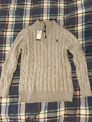 Ljusgrå kabelstickad tröja Polo Ralph Lauren - Säljer en ljusgrå kabelstickad tröja från Polo Ralph Lauren med half zip och broderad logga på bröstet. Helt oanvänd, prislappen sitter fortfarande kvar. 