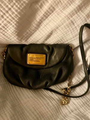 Marc by Marc Jacobs clutch - Säljer en svart clutch/axelväska från Marc by Marc Jacobs i mjukt skinn. Väskan har guldfärgade metalldetaljer, kedja och en ikonisk metallbricka framtill. Invändigt finns ett coolt mönstrat foder med logga och flera fack för kort och småsaker. I använt men fortfarande fint skick🩷