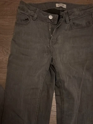 Grå jeans från Lindex  - Säljer ett par svarta jeans från Lindex i storlek 152. Klassisk femficksmodell med dragkedja och knapp framtill. Jeansen har utsvängda ben och bälteshällor, perfekta till vardagslooken. Materialet är mjukt och bekvämt bomullstyg.